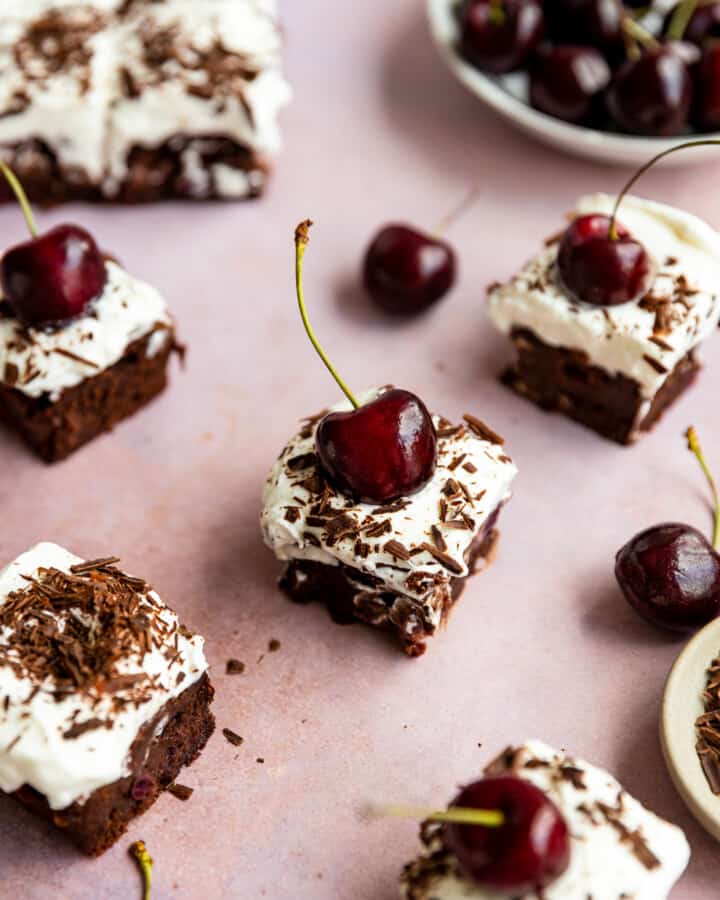 A few black forest brownies topped with whipped cream, shaved chocolate and a fresh cherrie on a pink surface.