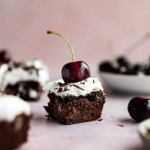 A close up of a black forest brownie on a table with cherries in the background.