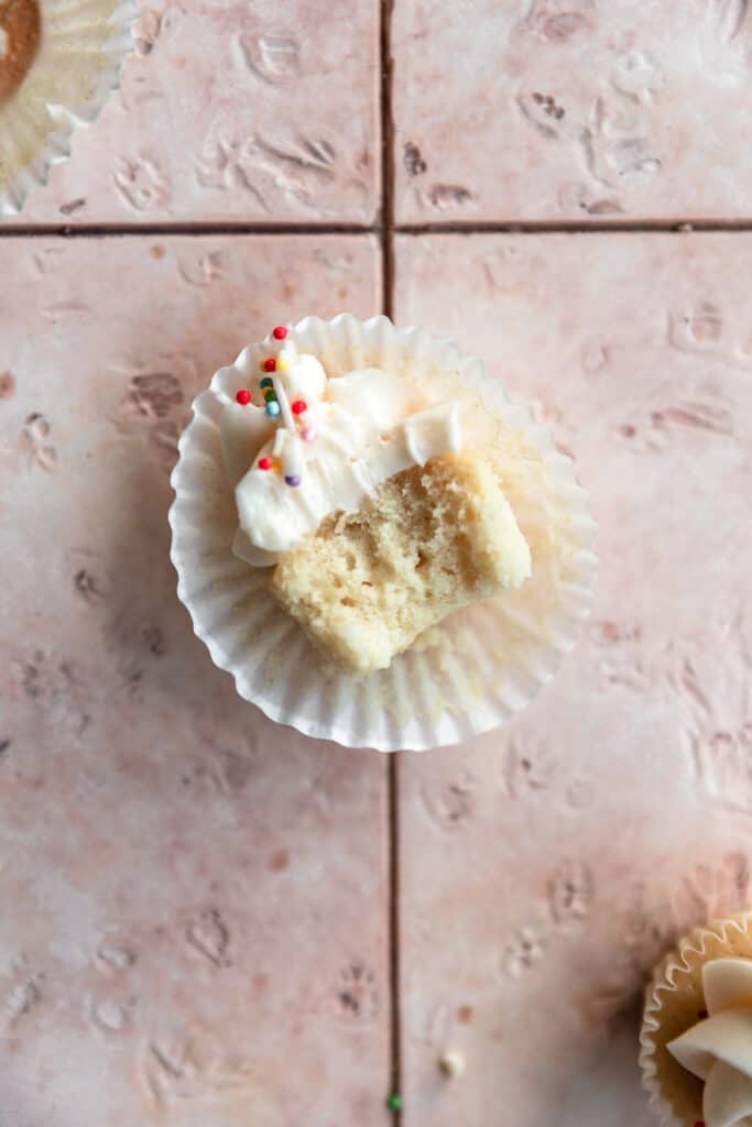 A mini vanilla cupcake with a bite taken out of it on a pink tile surface.