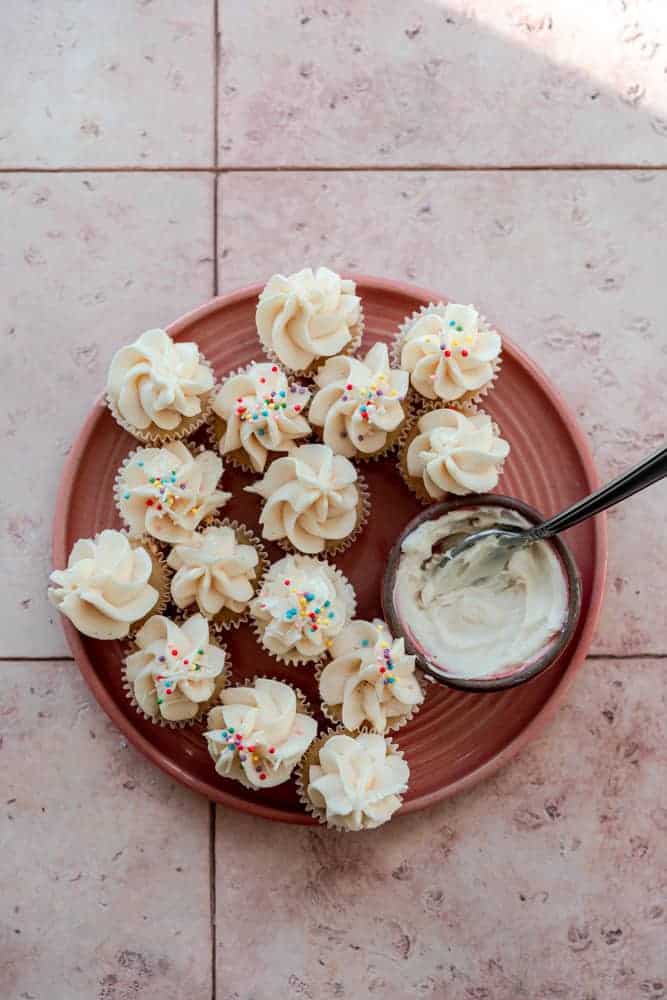 Mini cupcakes on a pink plate next to a bowl of vanilla frosting. 