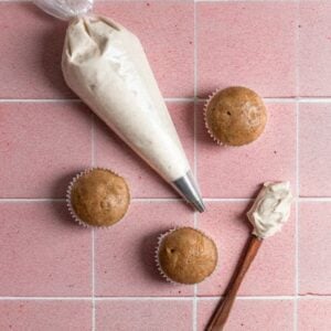 A piping bag with frosting next to un-frosted cupcakes on a pink tiled surface.