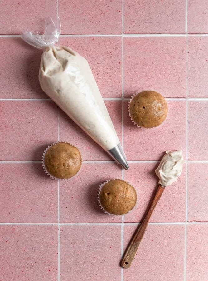 A piping bag with frosting next to un-frosted cupcakes on a pink tiled surface.