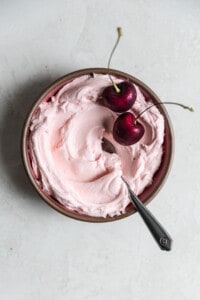 Pink cherry frosting in a small bowl with 2 fresh berries on top.