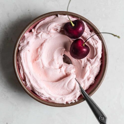 Pink cherry frosting in a small bowl with 2 fresh berries on top.