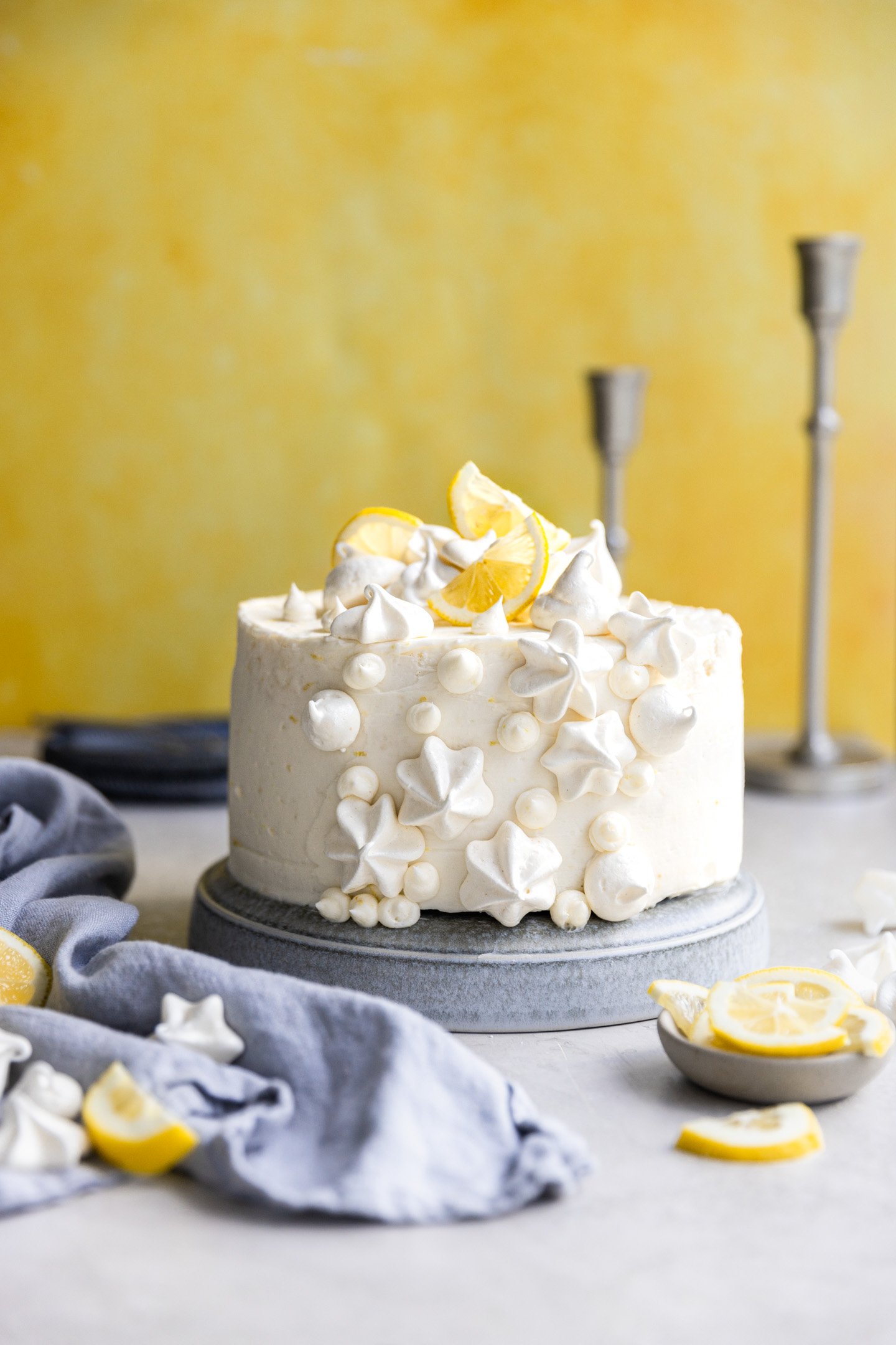 A lemon curd cake decorated with mini meringues and lemon slices on a light blue plate with a yellow background. 