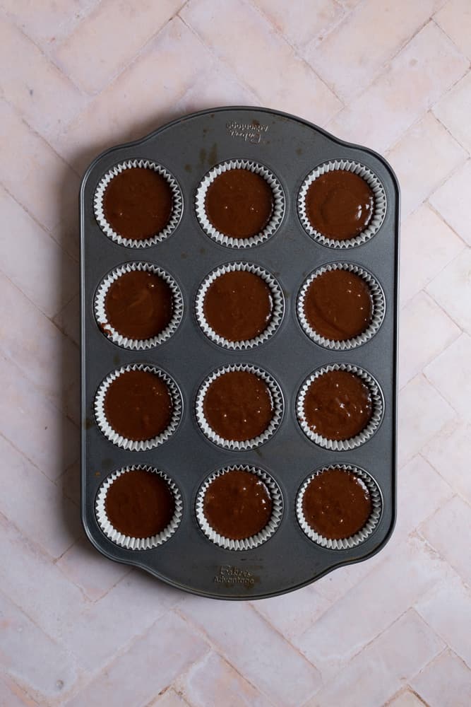Chocolate batter in a muffin tin on a pink tiled surface.