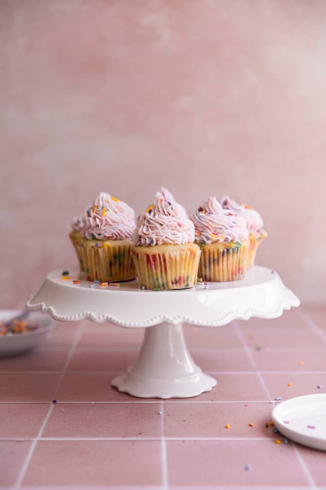 Funfetti cupcakes on a white cake stand on a pink surface.
