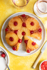 An upside down pineapple cake with one slice cut out on a white plate and yellow surface.