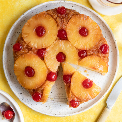An upside down pineapple cake with one slice cut out on a white plate and yellow surface.