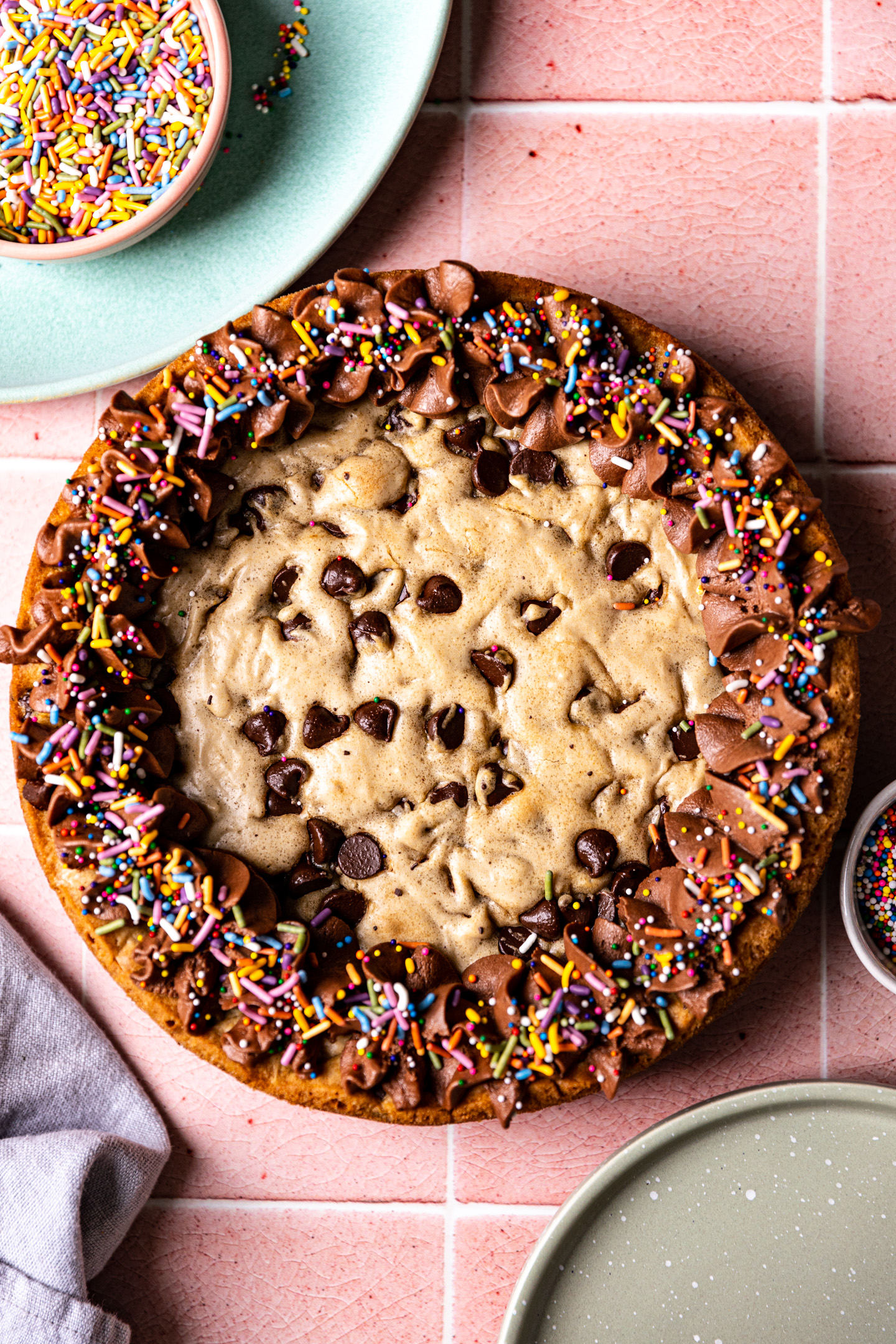 A chocolate chip cookie cake decorated with chocolate frosting and rainbow sprinkles on a pink tile surface.