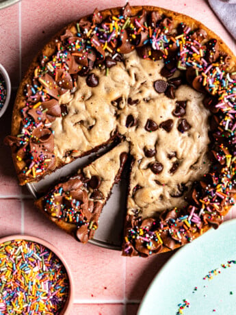 A chocolate chip cookie cake with chocolate frosting and sprinkles with a slice cut out on a pink background.