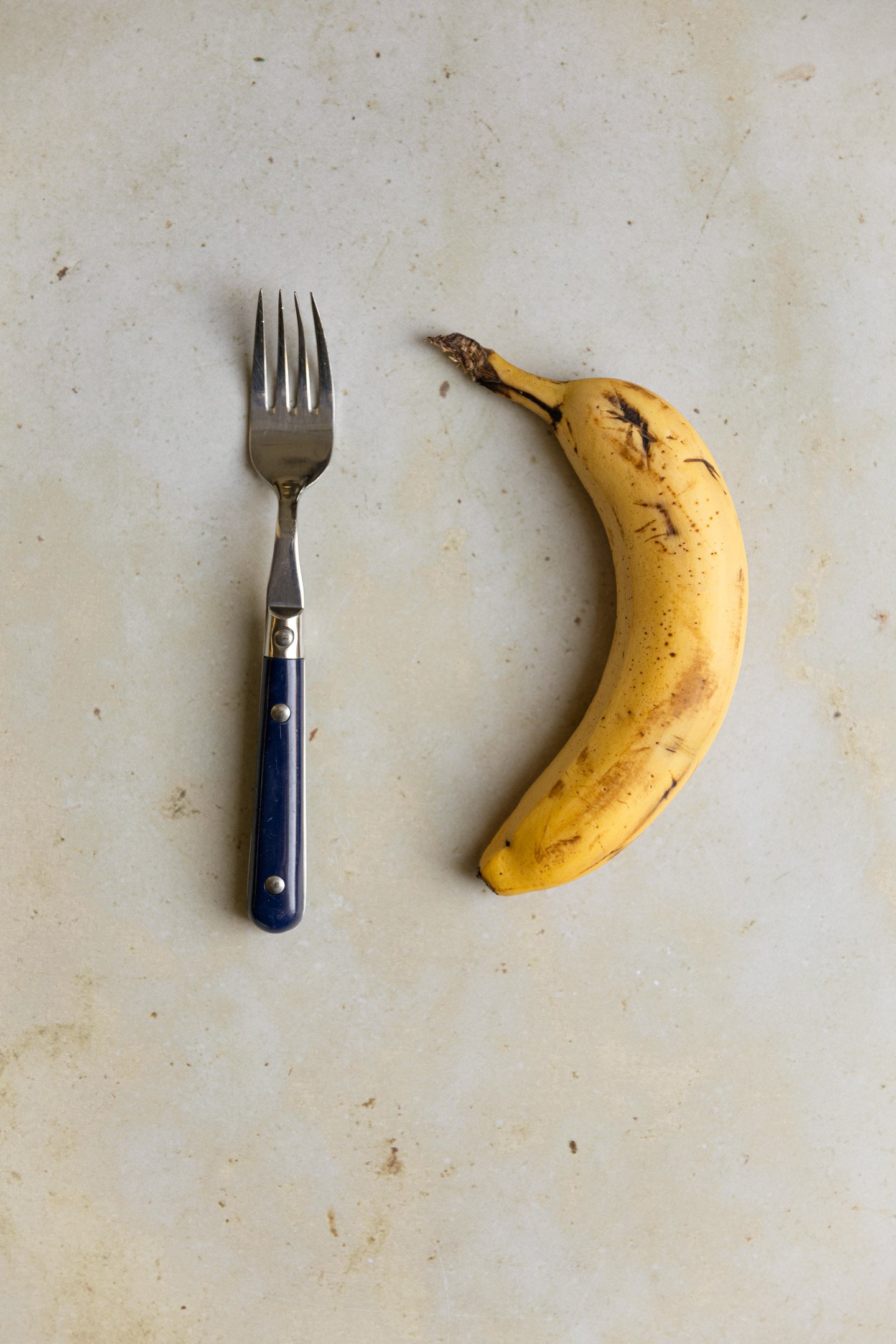 A banana next to a kitchen fork on a beige surface.