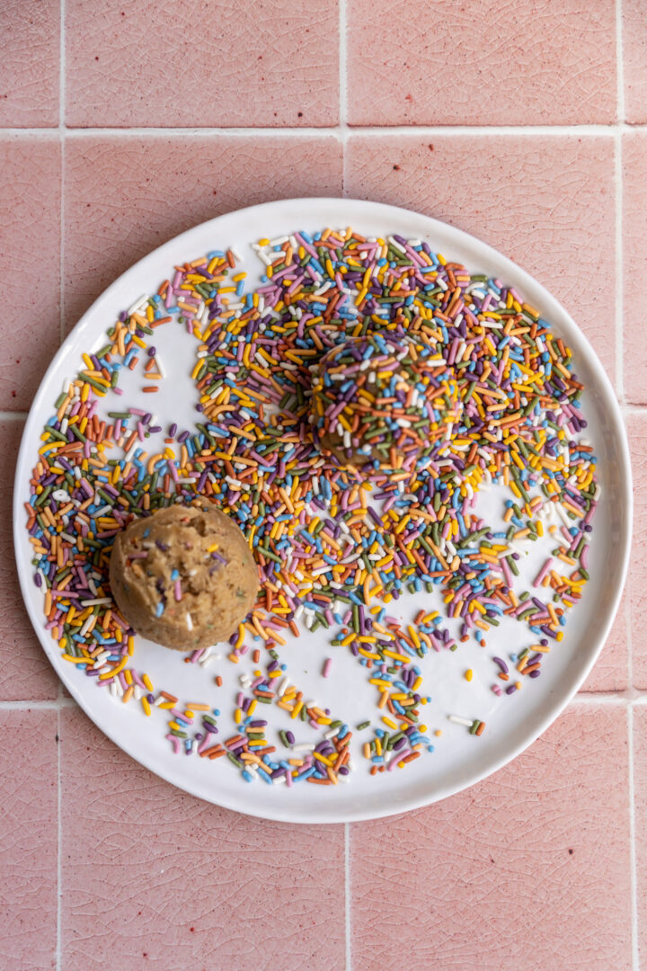 Cookie dough balls rolled in colorful sprinkles on a white plate.
