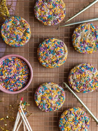 Birthday cake cookies on a cooling rack next to a bowl of sprinkles, candles, and matches.