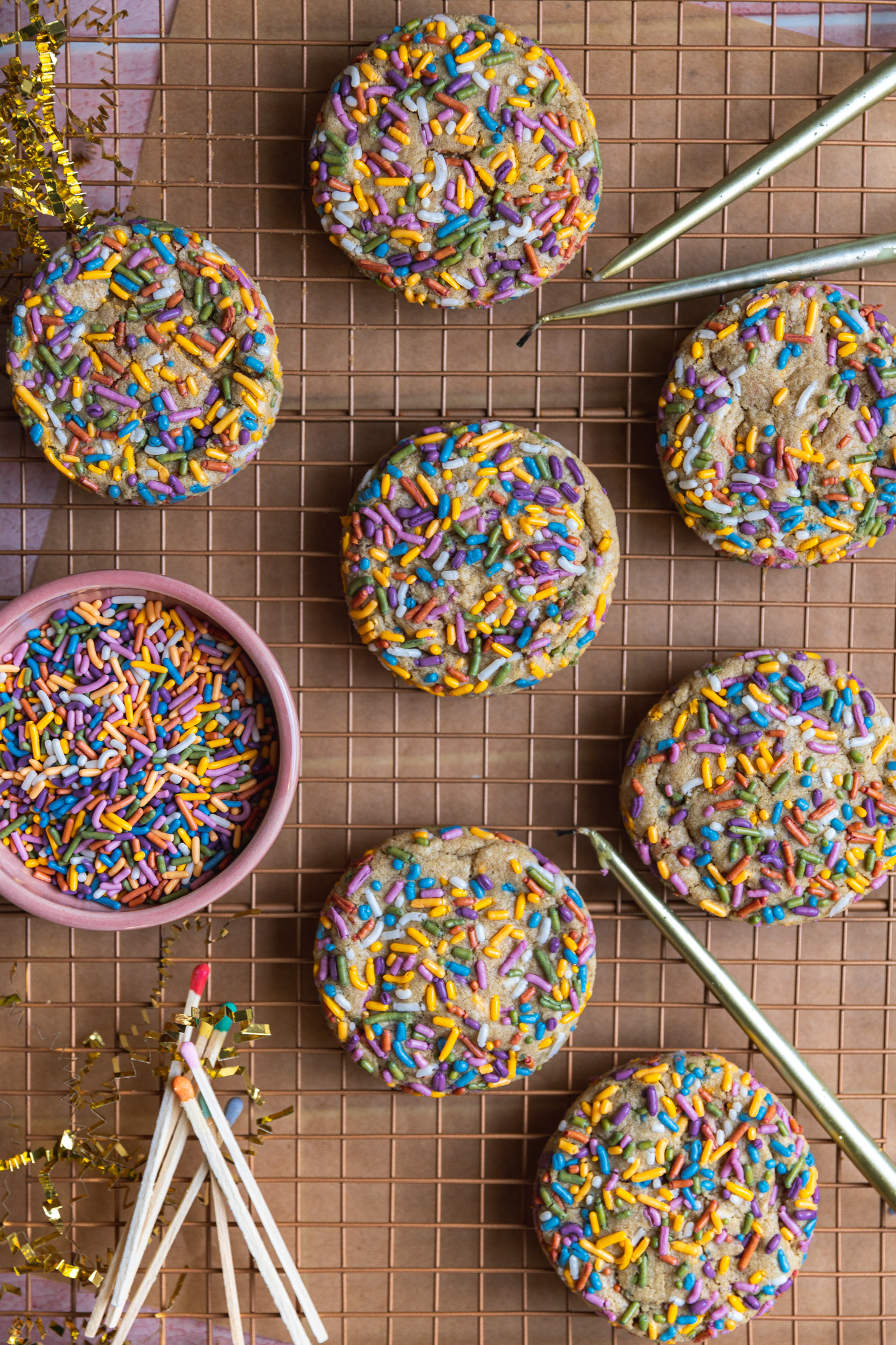Birthday cake cookies on a cooling rack next to a bowl of sprinkles, candles, and matches.