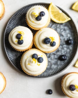 Four lemon blueberry cupcakes on a blue tinted plate with a slice of lemon and blueberries next to them.