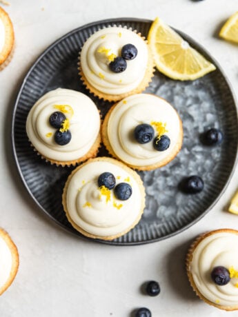 Four lemon blueberry cupcakes on a blue tinted plate with a slice of lemon and blueberries next to them.
