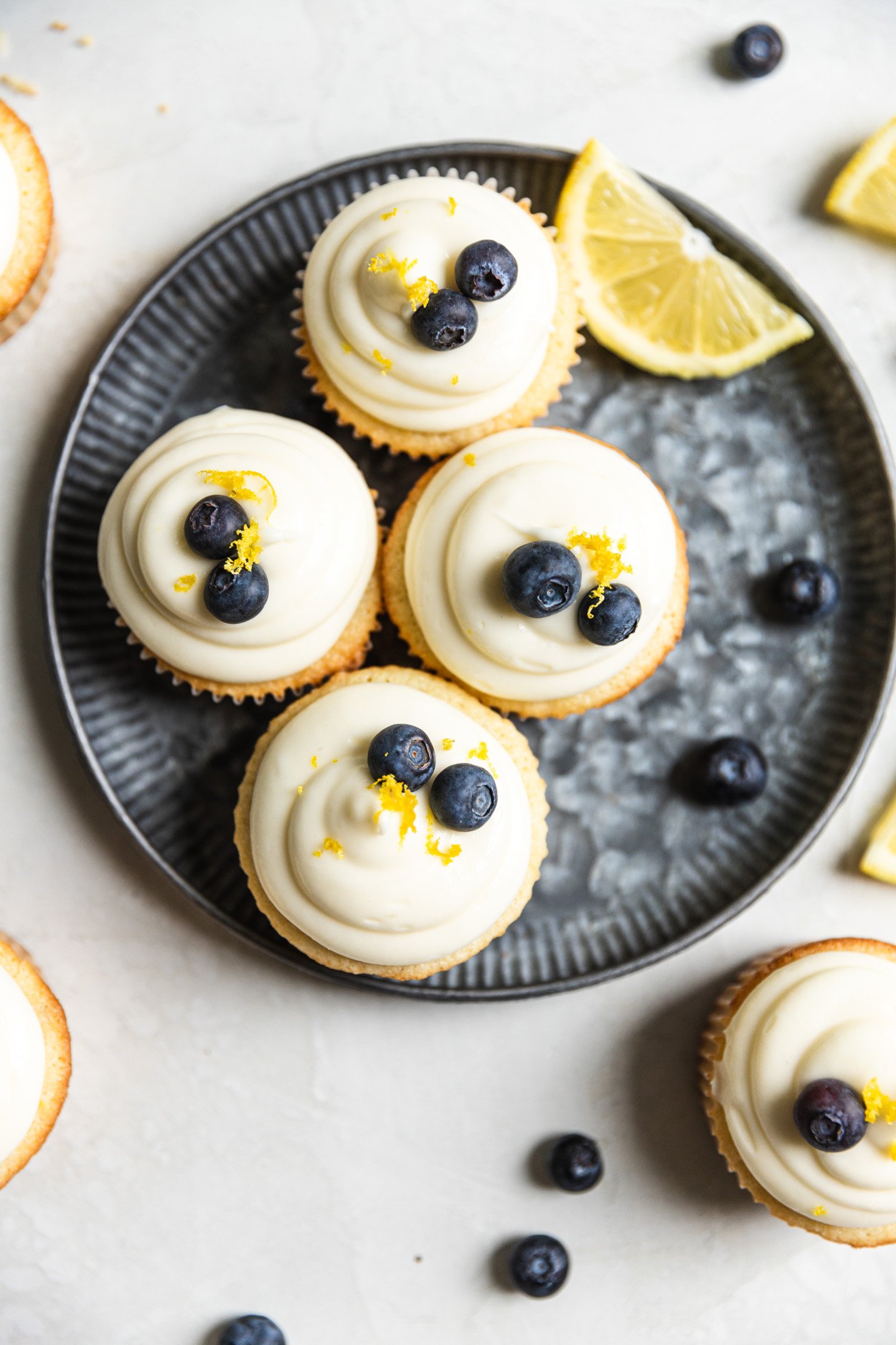 Four lemon blueberry cupcakes on a blue tinted plate with a slice of lemon and blueberries next to them. 