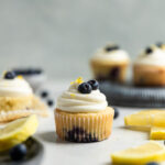 A lemon blueberry cupcake frosted with white frosting on a table next to other frosted cupcakes and slices of lemons.