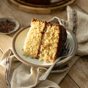 A slice of yellow cake with chocolate frosting on a rustic plate and linen napkin.