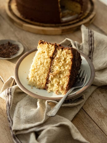 A slice of yellow cake with chocolate frosting on a rustic plate and linen napkin.