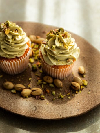 2 vanilla cupcakes with pistachio frosting on a brown platter surrounded by pistachios.