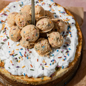 A birthday cake cheesecake topped with whipped cream and edible sugar cookie dough.