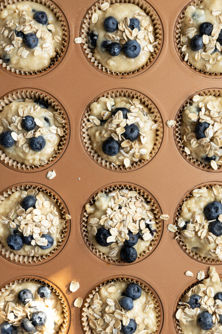 A muffin pan filled with batter and topped with blueberries and oats.