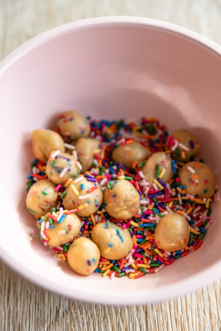 Mini balls of sugar cookie dough mixed in bowl of sprinkles.